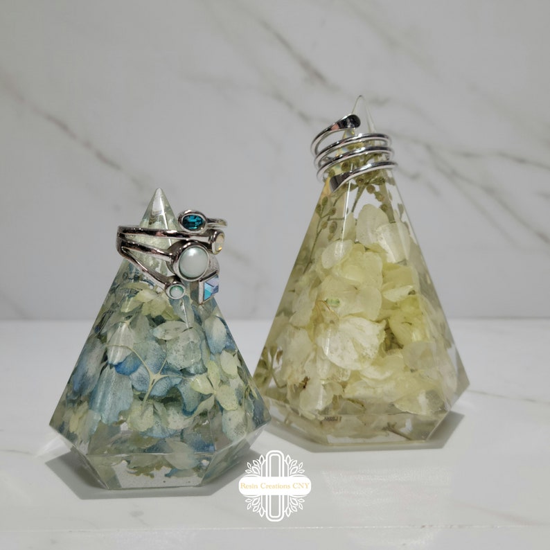 May include: Two clear resin ring holders shaped like pyramids. The holders are filled with dried flowers. One holder has white flowers and the other has blue and white flowers. The holders are on a white surface.