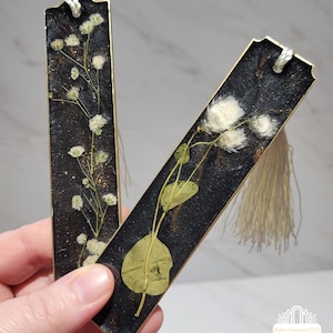 May include: Two rectangular resin bookmarks with gold edges. One features white flowers and the other has green leaves and white flowers, both set against a black background with gold flecks. Each bookmark has a tassel and a white cord.
