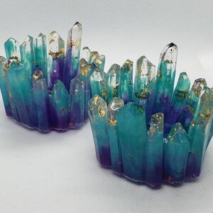 May include: Two teal and purple crystal candle holders with gold flecks. The holders are made of resin and are shaped like clusters of crystals.