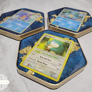 May include: Three hexagonal coasters made of blue resin with gold leaf flakes embedded inside. Each coaster features a different embedded Pokémon trading card, including Snorlax, Squirtle, and Blastoise, with gold-painted edges on the sides.