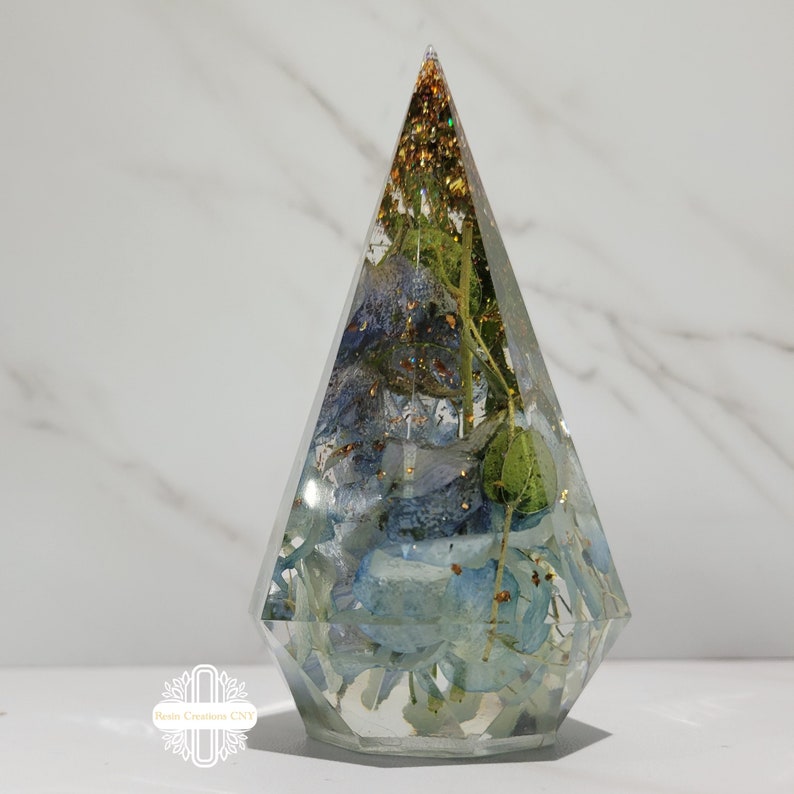 May include: A clear resin pyramid with a geometric shape, filled with dried blue and green flowers and gold glitter.
