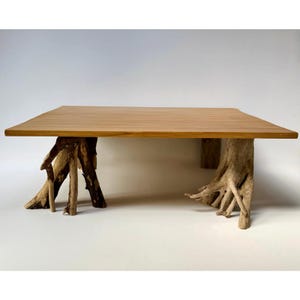 May include: A rectangular coffee table featuring a smooth, light-toned wooden tabletop surface supported by two distinct, weathered tree root bases. The natural, textured, and gnarled wood of the roots provides a contrasting foundation to the flat top.