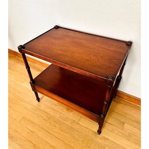 May include: A rectangular two-tier side table crafted from dark brown wood. The table features a smooth top surface and a lower shelf, supported by four vertical legs with decorative joints at each corner of the top frame and shelf levels.