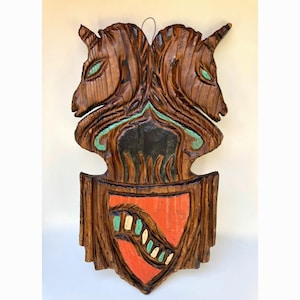 May include: A carved wooden wall hanging featuring two unicorn heads with green eyes and a shield. The shield is red with a white and green design. The wood has a natural brown finish.