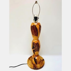 May include: A table lamp base crafted from polished wood with a natural grain pattern featuring light tan and dark brown swirls. The lamp has a stacked, rounded silhouette, a circular base, and a metal harp fixture at the top for a lampshade.