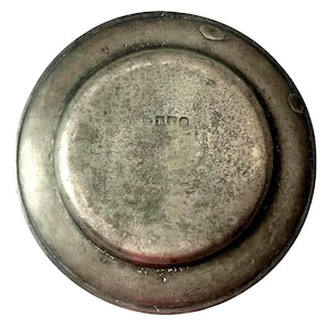May include: A vintage, round, silver-colored plate with a slightly raised rim. The plate's surface shows signs of age and wear, with a textured appearance. The center of the plate has a smaller, recessed area. The back of the plate has markings.