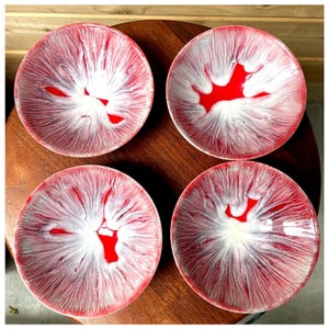 May include: Four red and white decorative plates with a unique, abstract design. Each plate features a central red shape surrounded by white and red radial patterns, creating a striking visual effect. The plates are arranged on a dark wooden surface.