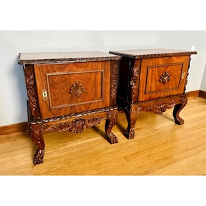 May include: A pair of matching dark brown wooden nightstands featuring a single cabinet door with a central carved shell motif, ornate floral carvings along the legs and edges, and a brass keyhole escutcheon on each door front.