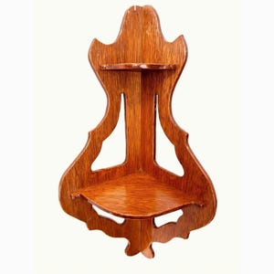 May include: A vintage, corner-mounted wooden shelf with a warm, reddish-brown finish. The shelf features two triangular platforms and an ornate, decorative back with curved edges and cut-out details. The shelf is designed to fit into a corner.