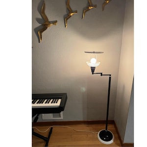 May include: A black and white floor lamp with a round base and a pivoting arm. The lamp has a frosted glass shade and a light bulb. The background includes a piano and gold bird wall decor.