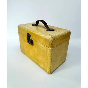 May include: A rectangular vanity case in a pale yellow hue with a textured, crackled surface finish. The case features a dark brown top handle and a central metal latch mechanism on the front panel for secure closure of the hinged lid.