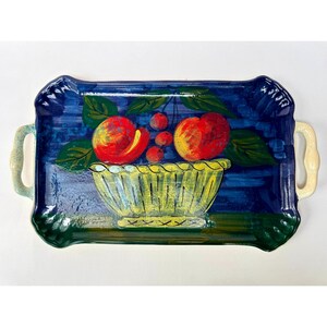 May include: A rectangular ceramic serving tray with two side handles, featuring a hand-painted design of a yellow bowl filled with red peaches and cherries against a deep blue background, with green leaves surrounding the fruit arrangement.
