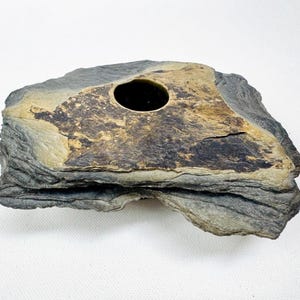 May include: A natural stone vessel with a layered, slate-like texture and a dark gray exterior. The top surface features a flat, tan-colored section with a circular opening in the center, revealing a dark, hollow interior space within the rock.