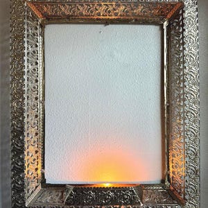 May include: Ornate silver-colored picture frame with intricate filigree detailing. The frame has a rectangular shape with a light emanating from within, creating a warm, golden glow. The frame is plugged into an electrical outlet.