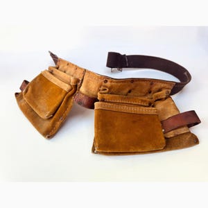 May include: A brown leather tool belt with multiple pockets and a brown elastic belt. The belt has a worn appearance with visible stitching and rivets. The pockets are various sizes, designed for carrying tools.