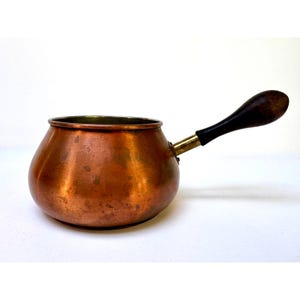 May include: A copper saucepan with a rounded body and a wooden handle. The handle is attached with a brass fitting. The pan has a dark, aged patina, suggesting it is vintage or antique. The interior appears to be a darker color, possibly from use.