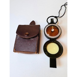 May include: A vintage compass with a brown leather case. The compass has a black frame, a circular dial with directional markings, and a needle. The leather case is rectangular with a flap and a button closure. A black cord is attached to the compass.