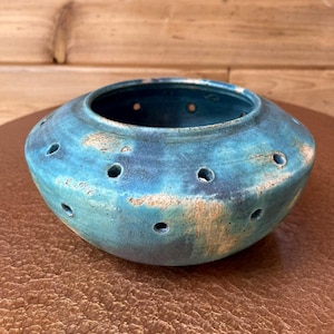 May include: A turquoise ceramic bowl with a series of evenly spaced holes around the sides. The bowl has a rounded shape with a wide opening and a textured surface. The color is a blend of blue and green with lighter, tan-colored accents.
