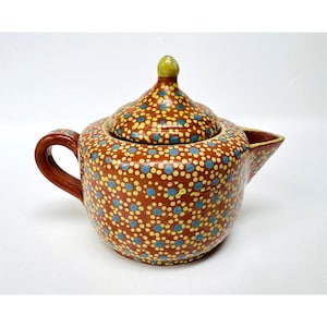 May include: A terracotta-colored ceramic teapot featuring a hand-painted pattern of small light blue and yellow dots. The teapot includes a matching lid with a small yellow knob, a curved handle on the left side, and a short, wide spout on the right.