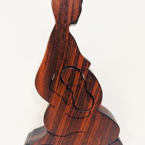 May include: A wooden sculpture of a pregnant person, with a cutout of a baby in the belly. The sculpture is made of dark brown wood and sits on a rectangular base. The wood grain is visible.