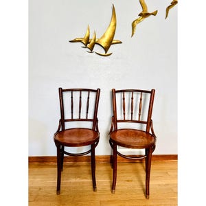 May include: Two matching wooden chairs with a dark brown finish, featuring rounded seats and backrests with three vertical spindles. The chairs have curved frames and four legs, each capped with a metallic detail at the bottom of the feet.