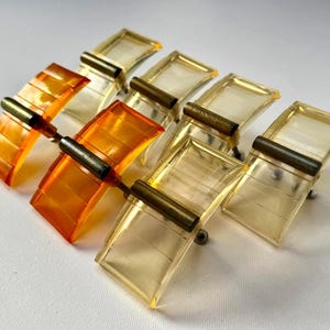 May include: A collection of eight vintage cabinet pulls. The pulls feature a rectangular, curved design with a transparent amber and yellow color. Each pull has a brass-colored metal bar and a small silver-colored screw.