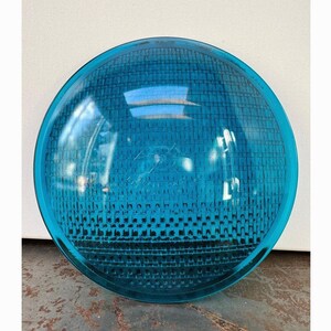 May include: A round, transparent, teal-colored glass object with a grid pattern. The object has a slightly domed shape and reflects light, creating a shimmering effect. The object is likely a light lens or cover.