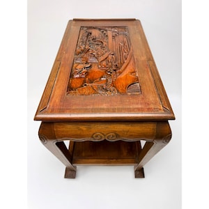 May include: A rectangular, wooden side table with a detailed, carved top featuring a scene with figures and architectural elements. The table has a lower shelf and curved legs, with decorative carvings on the apron. The wood has a warm, brown tone.