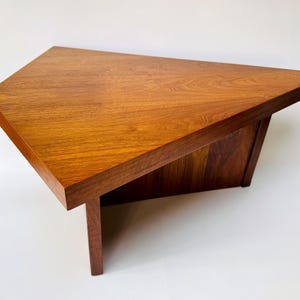 May include: A triangular wooden side table featuring a warm, medium-brown wood grain finish. The table has a flat, wide top surface and is supported by a solid, angular base structure that mirrors the triangular shape of the tabletop.