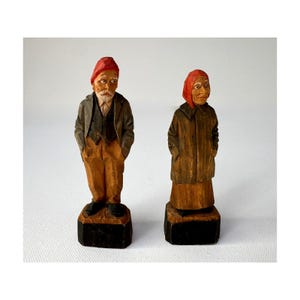 May include: Two hand-carved wooden figurines of an elderly couple. The man wears a red cap, jacket, and brown pants. The woman wears a red head covering and a long brown coat. Both figures stand on black rectangular bases.