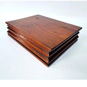 May include: A rectangular wooden box with a dark brown finish and visible wood grain. The box features a tiered, stepped edge design along the sides and two small, thin metal hinges attached to the top lid, allowing it to open and close securely.