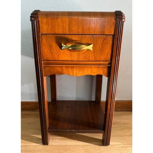 May include: A vintage wooden nightstand with a single drawer and a lower shelf. The nightstand has a warm, reddish-brown finish with a decorative brass-colored handle on the drawer. The design features curved edges and vertical grooves on the legs, showcasing an Art Deco style.