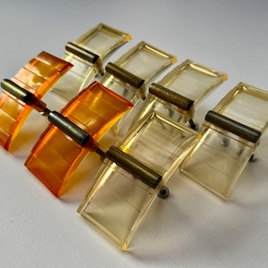May include: Set of eight vintage cabinet pulls. The pulls feature a rectangular shape with a curved design, made of amber and clear acrylic. Each pull has a brass-colored metal bar and a silver-colored screw.