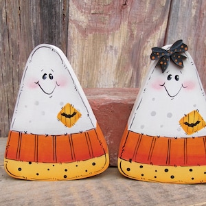 May include: Two triangular candy corn figures with painted faces, rosy cheeks, and stitched details. Each features a white top, orange middle, and yellow bottom with black dots. One figure has a black bow with orange polka dots on top.