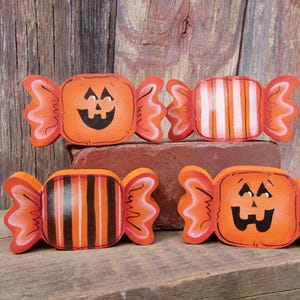 May include: Four wooden candy-shaped decorations featuring orange and black colors. Two pieces display painted jack-o'-lantern faces with black eyes and mouths, while the other two pieces feature vertical striped patterns in orange, black, and white.