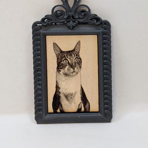 May include: A framed cat portrait. The black frame has an ornate, heart-shaped top and a textured border. The cat portrait is a black and white drawing on a tan background. The cat has large eyes and is looking forward.