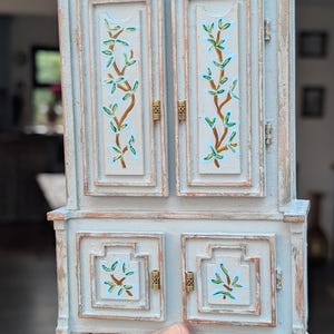 以下が含まれることがあります： 上部に2つのドア、下部に2つのドアが付いた、淡い青色のミニチュアキャビネット。ドアは茶色、緑、青の花柄で装飾されています。キャビネットは金色の金具と装飾的な足が付いています。高さ約15cm。