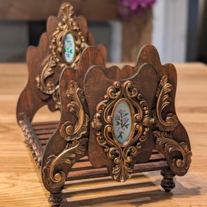 May include: A dark brown wooden decorative bed-shaped object with gold-colored ornate detailing. The object features an oval-shaped inset with a floral design. The bed has a slatted base and decorative legs.