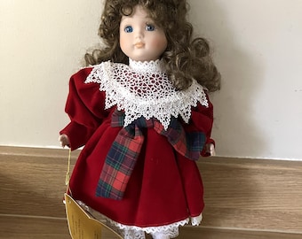 May include: A vintage porcelain doll with curly brown hair and blue eyes. The doll wears a red velvet dress with a white lace collar and a plaid bow. Black shoes complete the outfit. A tag hangs from the doll.