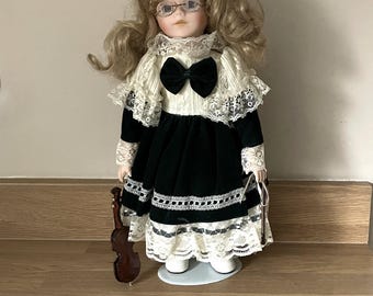 May include: A porcelain doll with curly blonde hair, wearing glasses, and a black velvet dress with white lace trim. The doll holds a miniature violin. The doll is standing on a white oval base.