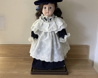 May include: A porcelain doll wearing a white dress with lace trim and a blue velvet hat. The doll has brown hair and blue eyes.