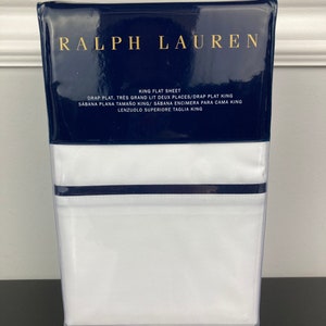 May include: A white king-sized flat sheet with a blue border, packaged in a clear plastic bag. The packaging features the Ralph Lauren logo and text in multiple languages indicating the sheet size and type.