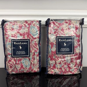 Op de afbeelding: Twee kussenslopen in transparante verpakking. De slopen hebben een bloemenpatroon in rood, blauw en cr&egrave;me. Elke verpakking heeft een label met het Ralph Lauren logo en tekst.