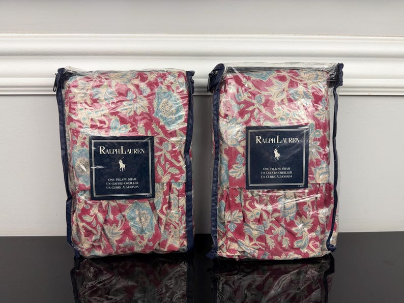 Op de afbeelding: Twee kussenslopen in transparante verpakking. De slopen hebben een bloemenpatroon in rood, blauw en cr&egrave;me. Elke verpakking heeft een label met het Ralph Lauren logo en tekst.