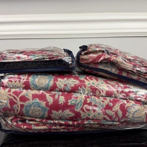 Op de afbeelding: Een beddengoedset, inclusief een dekbedovertrek en kussenslopen, is verpakt in doorzichtig plastic. Het beddengoed heeft een bloemenpatroon in rood, blauw en beige. De set is netjes opgevouwen en klaar voor gebruik.