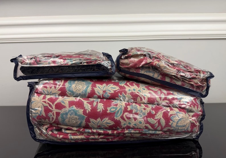 Op de afbeelding: Een beddengoedset, inclusief een dekbedovertrek en kussenslopen, is verpakt in doorzichtig plastic. Het beddengoed heeft een bloemenpatroon in rood, blauw en beige. De set is netjes opgevouwen en klaar voor gebruik.