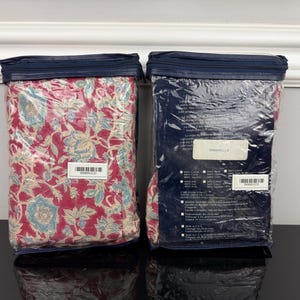Op de afbeelding: Twee verpakte beddengoedsets. E&eacute;n heeft een rood bloemenpatroon met blauwe en cr&egrave;mekleurige accenten, de andere zit in een marineblauwe verpakking. Beide zijn verzegeld in helder plastic met een marineblauwe ritssluiting.