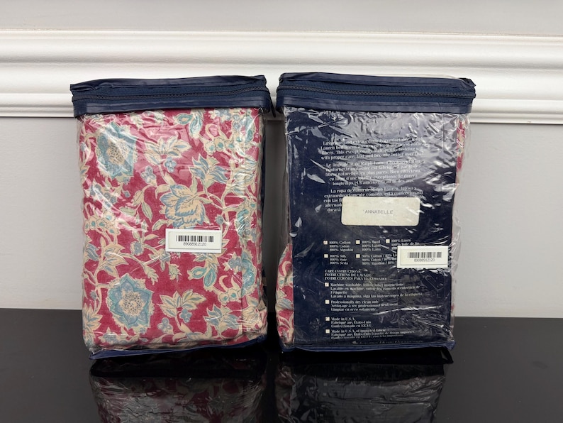 Op de afbeelding: Twee verpakte beddengoedsets. E&eacute;n heeft een rood bloemenpatroon met blauwe en cr&egrave;mekleurige accenten, de andere zit in een marineblauwe verpakking. Beide zijn verzegeld in helder plastic met een marineblauwe ritssluiting.
