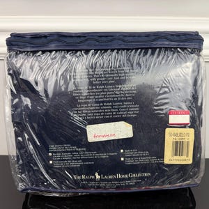 Op de afbeelding: Een marineblauwe beddengoedset, verpakt in doorzichtig plastic. De verpakking bevat tekst in meerdere talen, een "Clearance"-sticker en een label met een streepjescode. De set is van The Ralph Lauren Home Collection.
