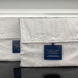 May include: Two folded, light gray bedding sheet sets with button closures. Each set has a dark blue tag with white text. The sheets have a subtle floral pattern. The sets are displayed on a black surface.